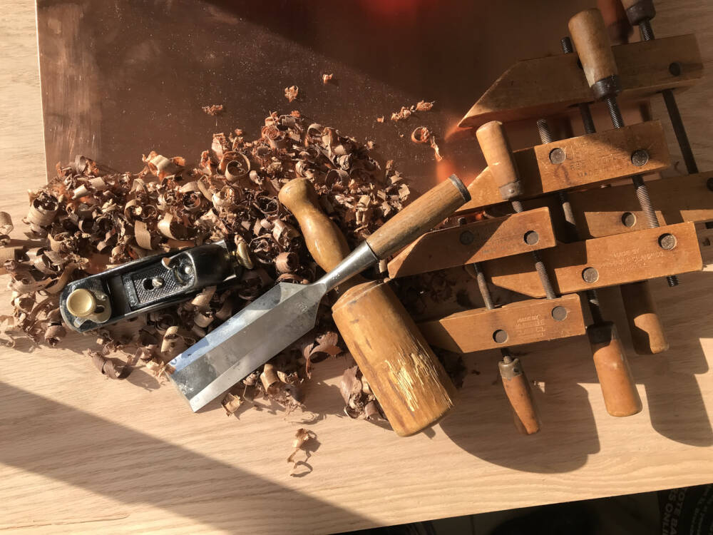 Carpentry, woodworking tools and wood shavings are assembled in the sunlight falling on a backdrop of red oak lumber and copper sheet metal.