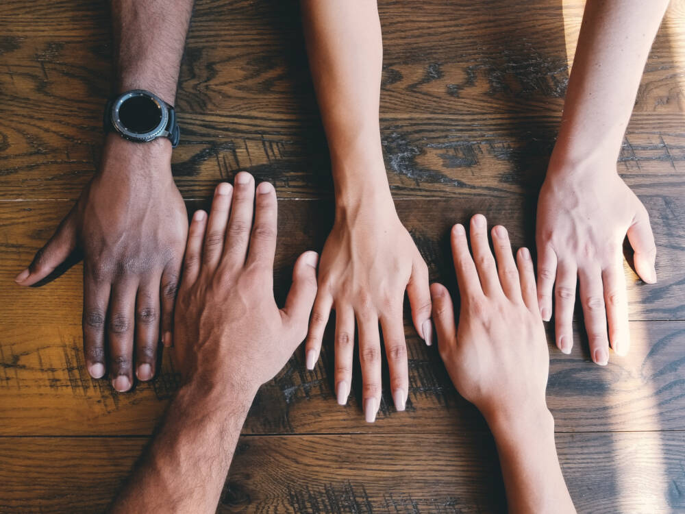 Hands of people of different ethnicities (as evidenced by skin colour) are laid on the background of a dark oak hardwood table to represent Classic Muskoka Homes' commitment to treating all people with equity.