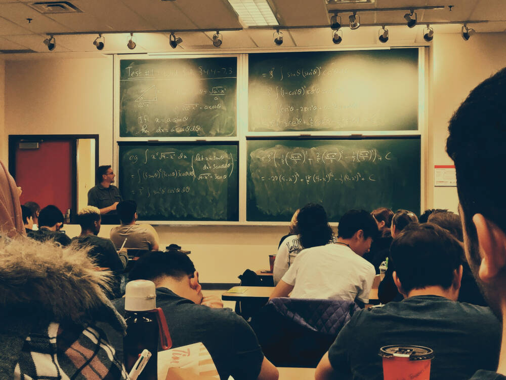 Students face a professor and green chalk board filled with math formulas and calculations to represent the topics of builder education, training, qualification, certification and licensing.