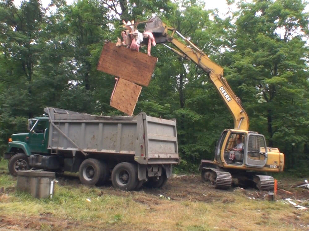 A demolition contractor uses a hydraulic excavator to demolish a whole wood frame building and load it into dump trucks to be hauled for disposal.