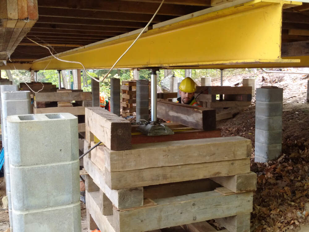 A building mover's staff install steel beams and wooden crib timbers to lift and temporarily support a wood frame house while its foundation is replaced in Muskoka, Ontario.