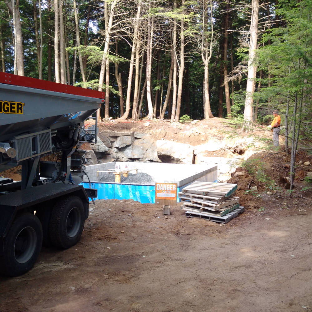 A slinger truck unloads gravel using a moving rubber conveyor belt to throw the material into place around a building foundation.