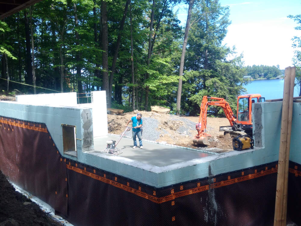 A large foundation construction project is busy with concrete finishing and excavation work while a radio plays pop music in the foreground on Lake Muskoka, Ontario.