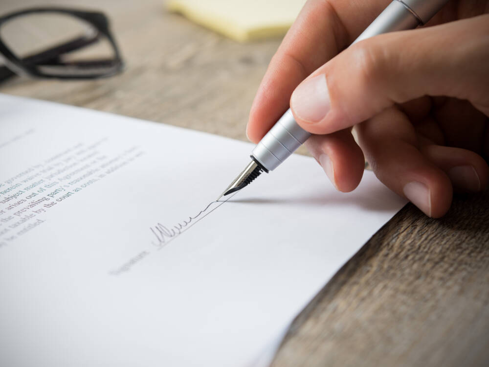 A hand holds an ink pen while adding the signature to a building construction contract document.
