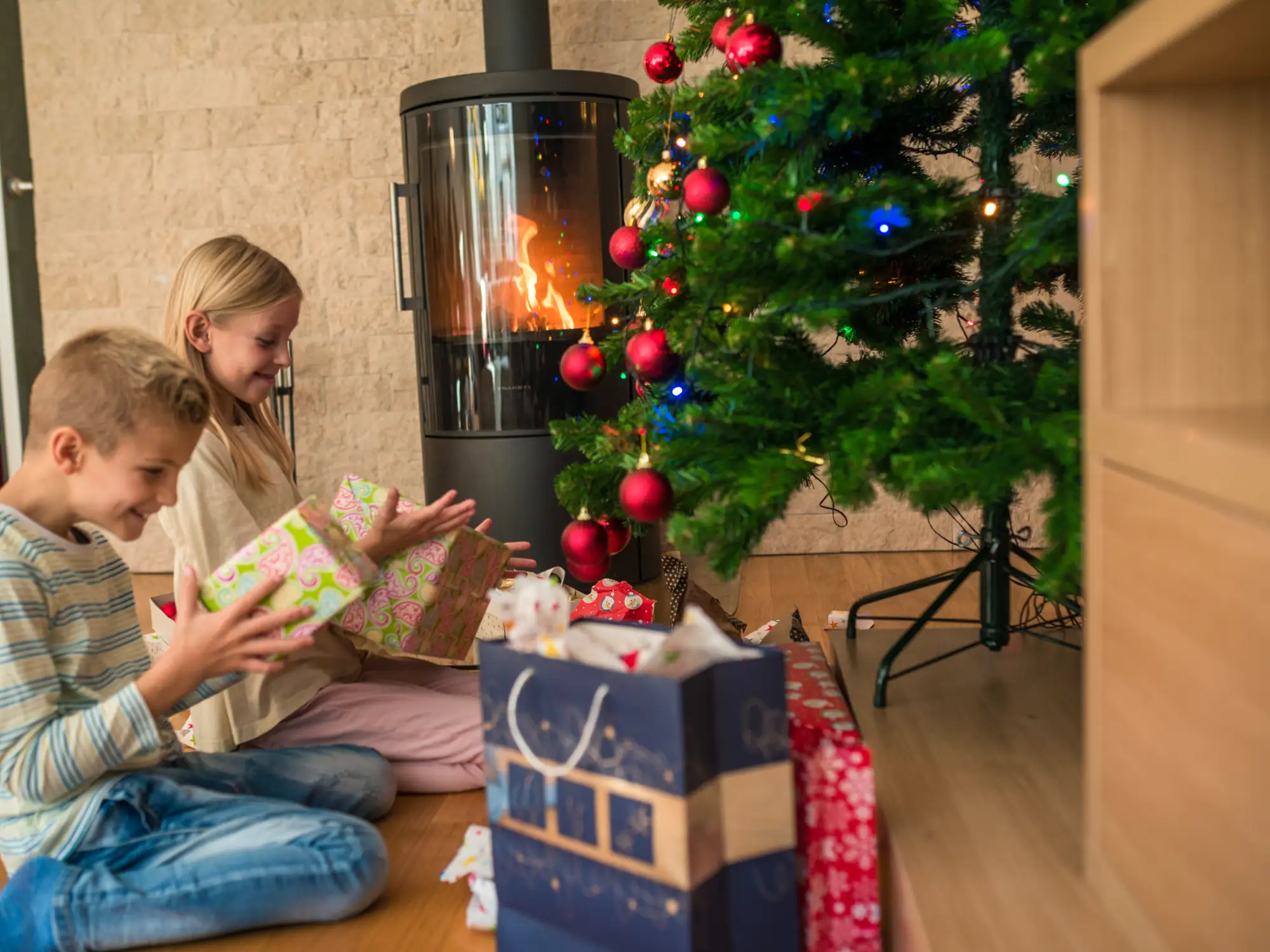 Young children open gifts on Christmas morning beside a woodstove in the living room of a family's average quality home.